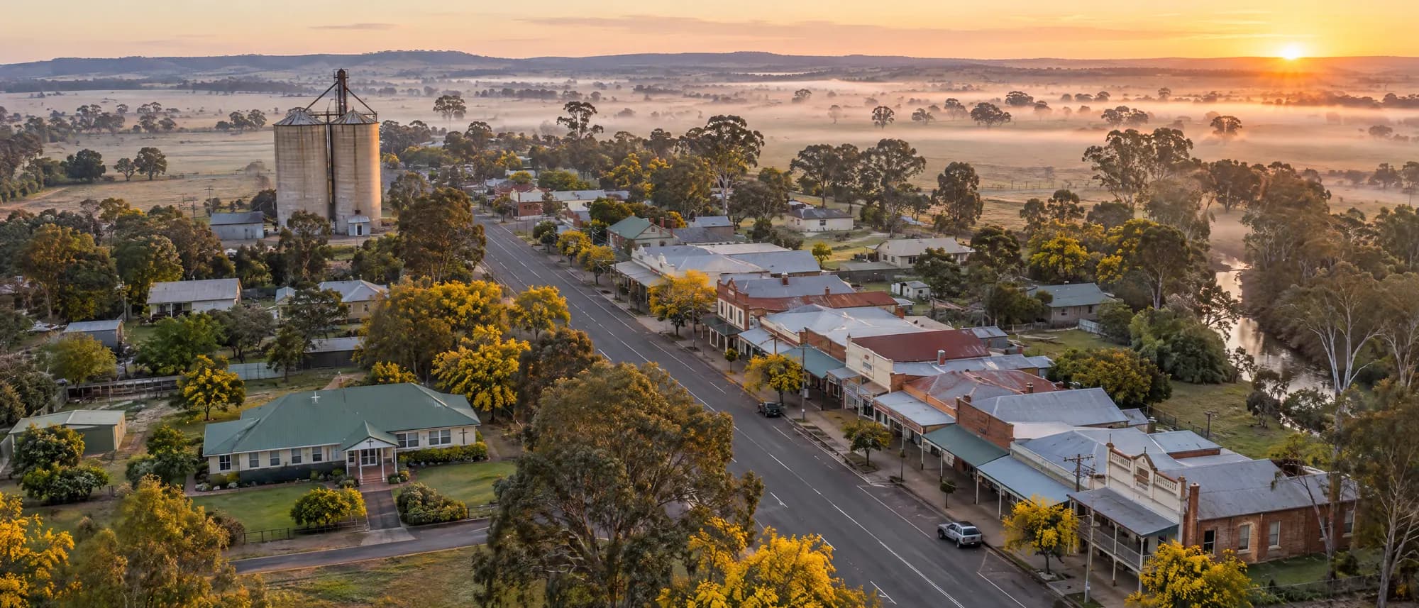 Aerial view of a small country town at dawn, with grain silos, a main street and a small hospital among gum trees.